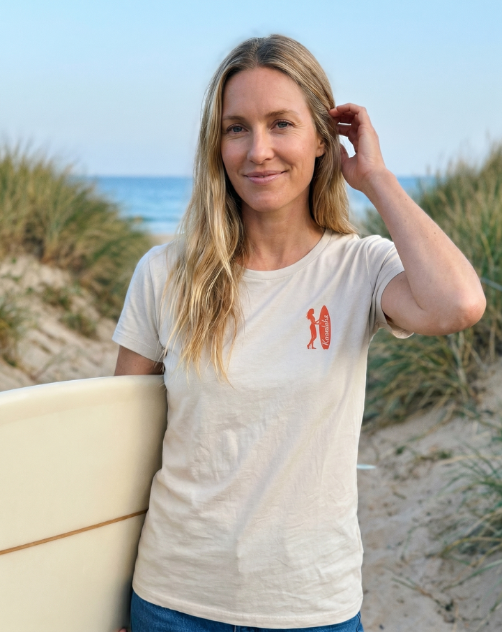Camiseta de bambú para mujer color marfil con diseño Nalu, modelo de 40 años en la playa con tabla de surf